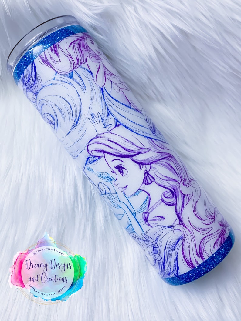 Puede incluir: Un vaso blanco con un borde de purpurina azul y un dibujo de contorno morado y azul de una princesa Disney con el pelo largo. El vaso tiene una pegatina con el texto "Dreamy Designs and Creations" en la parte inferior.