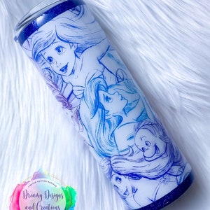 Puede incluir: Un vaso de acero inoxidable blanco con fondo de purpurina azul. El vaso presenta un dise&ntilde;o azul y blanco degradado con un boceto de Ariel y otros personajes de La Sirenita.