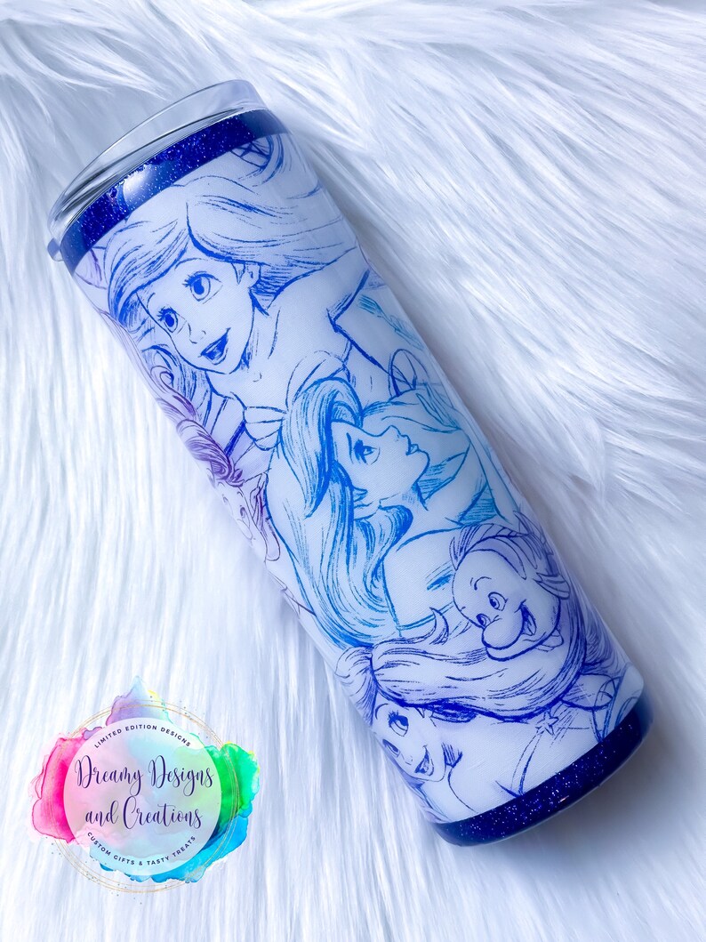 Puede incluir: Un vaso de acero inoxidable blanco con fondo de purpurina azul. El vaso presenta un dise&ntilde;o azul y blanco degradado con un boceto de Ariel y otros personajes de La Sirenita.
