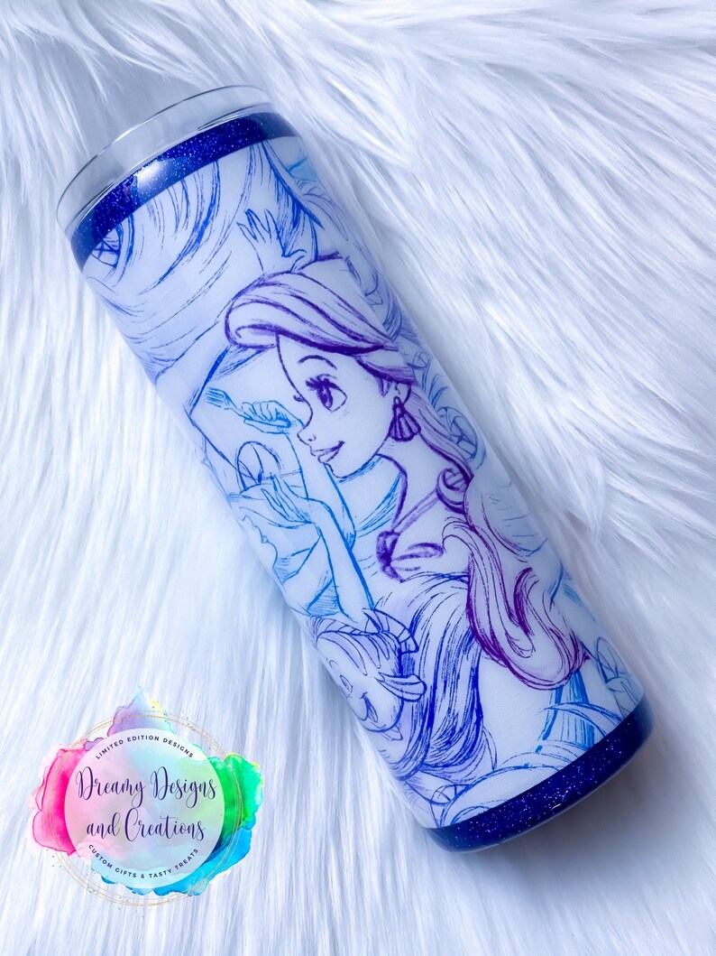 Puede incluir: Un vaso de acero inoxidable blanco con un borde de purpurina azul y un dibujo de l&iacute;nea azul y morado de una princesa con el pelo largo. El vaso tiene una pegatina con el texto "Dreamy Designs and Creations Custom Gifts & Tasty Treats".