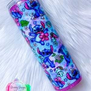 May include: A stainless steel tumbler with a pink glitter rim and a blue and white design featuring Stitch from Lilo & Stitch. The design includes Stitch in various poses, flowers, and other tropical elements. The tumbler is 20 ounces.