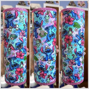 May include: A stainless steel tumbler with a pink glitter rim and a blue and white pattern featuring Stitch from Lilo & Stitch. The pattern includes flowers, leaves, and other tropical elements.