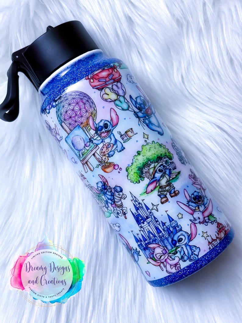Op de afbeelding: Een blauwe en witte waterfles van roestvrij staal met een zwarte dop en handvat. De fles is versierd met een patroon van Stitch-personages uit Disney's Lilo & Stitch, samen met verschillende Disney-parkiconen, waaronder Assepoester's kasteel, Spaceship Earth en de Tree of Life. De fles heeft een blauwe glitterafwerking.