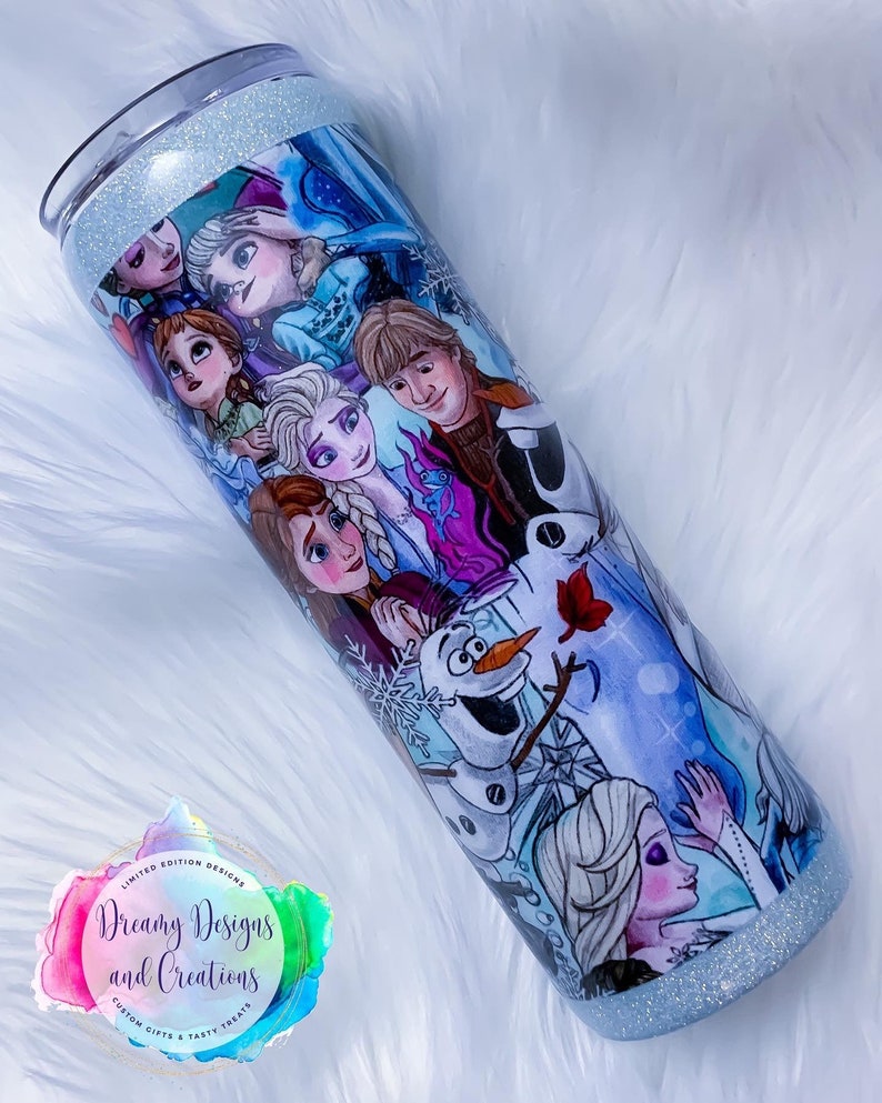 Puede incluir: Un vaso de acero inoxidable azul y blanco con un borde de purpurina. El vaso presenta un collage de personajes de la pel&iacute;cula animada Frozen, incluyendo Elsa, Anna, Olaf, Kristoff y Sven. El vaso tiene un fondo blanco con un borde de purpurina azul y blanco. El texto "Dreamy Designs and Creations" est&aacute; impreso en el vaso.