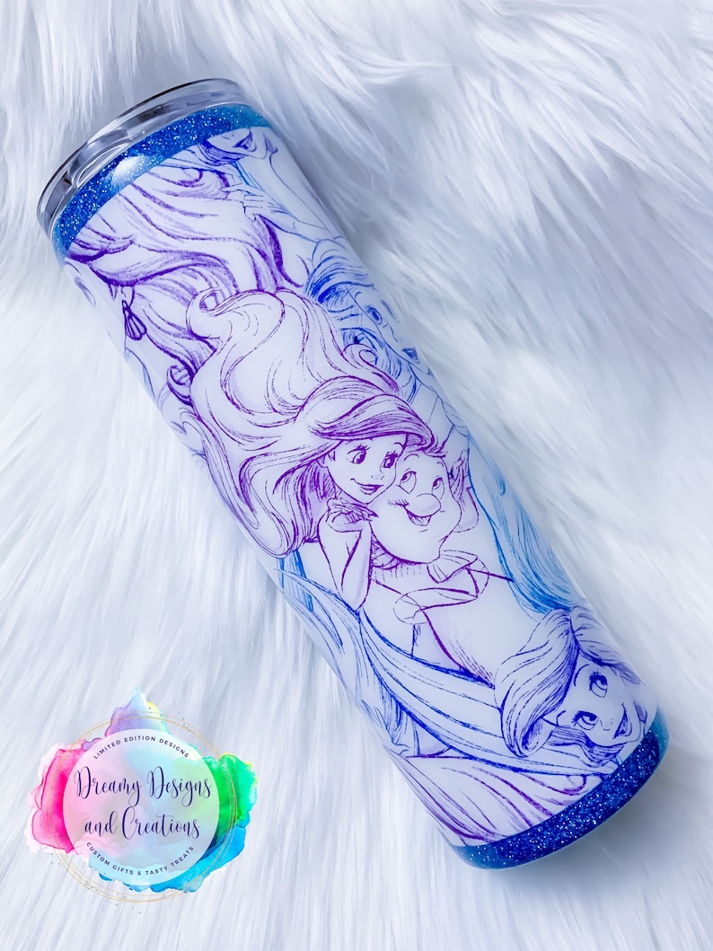 Puede incluir: Un vaso de acero inoxidable blanco con fondo de purpurina azul. El vaso presenta un dise&ntilde;o degradado de colores de varias princesas Disney, incluyendo Ariel, Cenicienta y Bella. El dise&ntilde;o es de estilo boceto y est&aacute; contorneado en morado, azul y rosa. El vaso tiene una tapa transparente y est&aacute; decorado con una pegatina que dice "Dreamy Designs and Creations".
