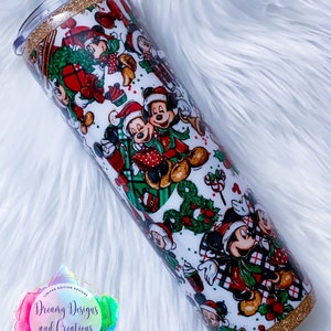 Pode incluir: Um copo branco com uma borda e base de glitter dourado. O copo apresenta um padr&atilde;o de Natal com Mickey Mouse e Minnie Mouse em trajes vermelhos e verdes. O texto "Dreamy Designs and Creations" &eacute; vis&iacute;vel em um adesivo.