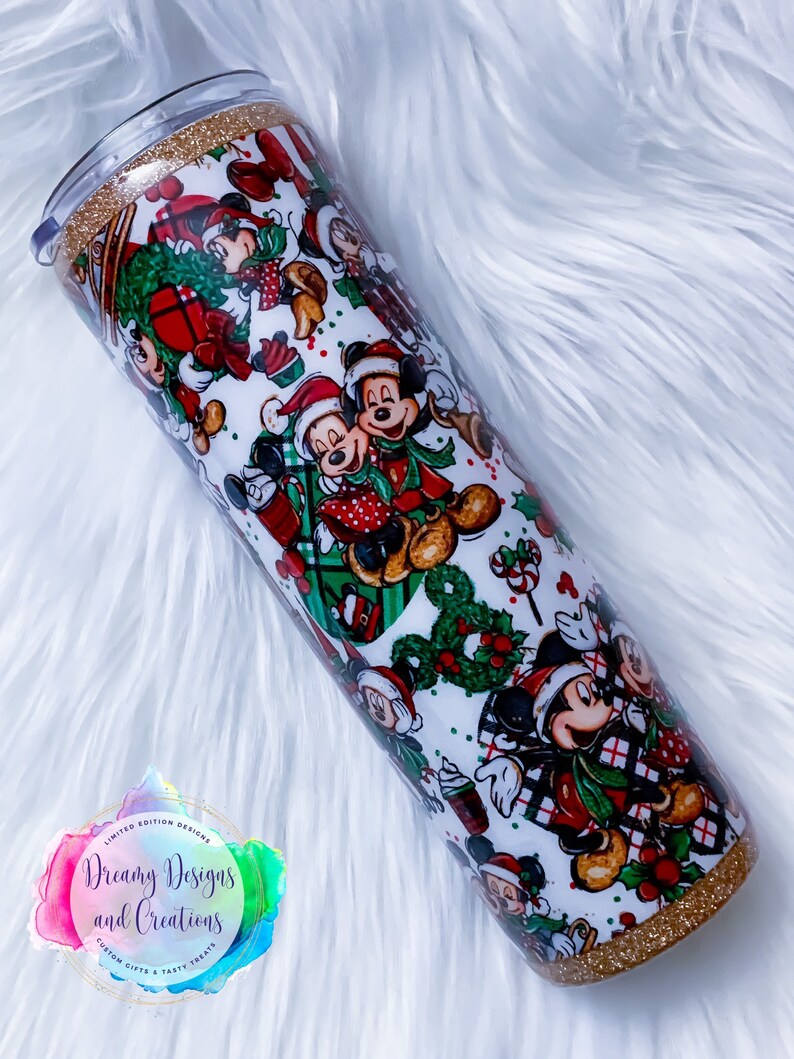 Pode incluir: Um copo branco com uma borda e base de glitter dourado. O copo apresenta um padr&atilde;o de Natal com Mickey Mouse e Minnie Mouse em trajes vermelhos e verdes. O texto "Dreamy Designs and Creations" &eacute; vis&iacute;vel em um adesivo.