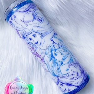Puede incluir: Un vaso blanco con fondo de purpurina azul. El vaso presenta un dise&ntilde;o degradado de colores con un boceto de los personajes de La Sirenita de Disney. El texto "Dreamy Designs and Creations" es visible en el vaso.
