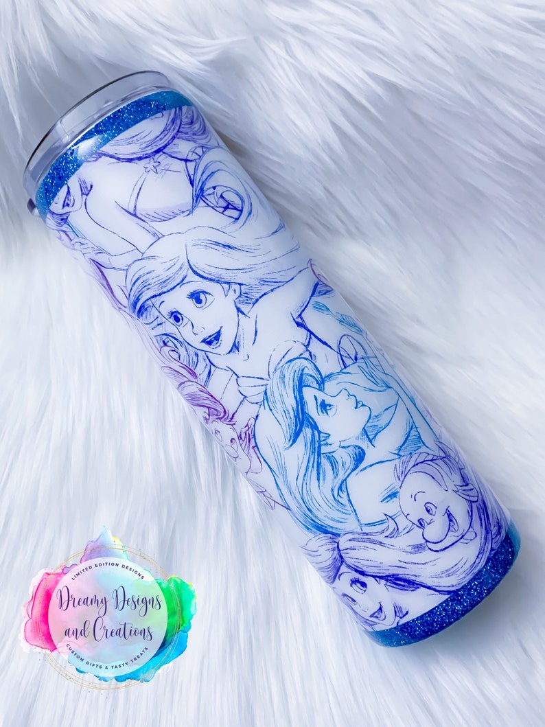 Puede incluir: Un vaso blanco con fondo de purpurina azul. El vaso presenta un dise&ntilde;o degradado de colores con un boceto de los personajes de La Sirenita de Disney. El texto "Dreamy Designs and Creations" es visible en el vaso.
