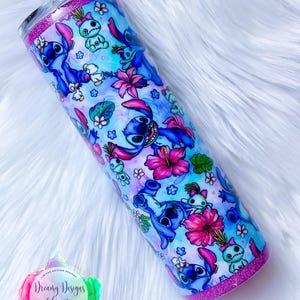 May include: A stainless steel tumbler with a pink glitter rim and a blue and white design featuring Stitch characters from the Disney movie Lilo & Stitch. The tumbler is decorated with flowers and a watercolor background.