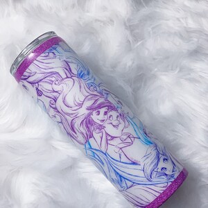 Puede incluir: Un vaso de acero inoxidable con un borde de purpurina morado y un fondo blanco con un dibujo de l&iacute;nea azul y morado de Ariel y el Pr&iacute;ncipe Eric de La Sirenita.