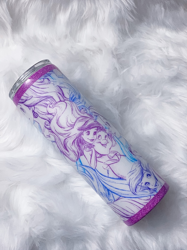 Puede incluir: Un vaso de acero inoxidable con un borde de purpurina morado y un fondo blanco con un dibujo de l&iacute;nea azul y morado de Ariel y el Pr&iacute;ncipe Eric de La Sirenita.