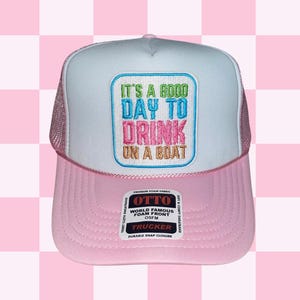 May include: A white and pink trucker hat with a mesh back. The front of the hat has an embroidered patch that says "It's a good day to drink on a boat". The hat also has a black label with the words "Otto World Famous Foam Front OSFM Trucker Durable Snap Closure" on it.