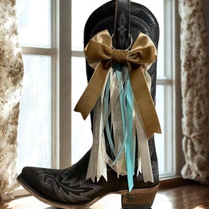 May include: Black leather cowboy boot with decorative stitching, adorned with a large tan bow and flowing ribbons in shades of tan, silver, and light blue. The boot is positioned near a window, suggesting a rustic or country theme.