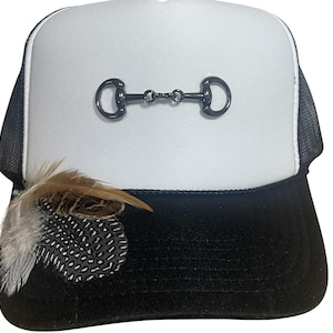 May include: Black and white trucker hat with a white front panel and black mesh back. The hat features a dark metal horse bit and decorative feathers on the brim. A stylish accessory for equestrian enthusiasts.