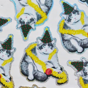 May include: Stickers of a grey and white cat wearing a Christmas tree hat and yellow garland. The cat is also holding a red ball. The stickers have a glittery, silver border.