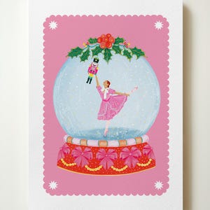 May include: A pink Christmas card featuring a snow globe with a ballerina holding a nutcracker doll. The snow globe is decorated with red bows and gold beads. The background is pink with white stars.
