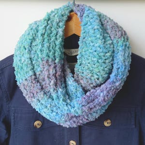May include: A blue and purple infinity scarf made of a soft, fuzzy yarn. The scarf is folded in half and draped over a blue jacket.