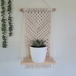 May include: A cream-coloured macrame wall hanging with a wooden shelf, displaying a potted succulent. The macrame features a geometric design and fringe. The shelf is made of light-coloured wood and is suspended by cream-coloured cords.