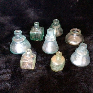 Op de afbeelding: Collectie van negen vintage glazen inktpotten in verschillende vormen en maten. De flessen zijn helder en hebben een lichtgroene tint. Sommige flessen hebben een verzilverde afwerking.