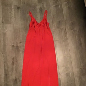 以下が含まれることがあります： 細いショルダーストラップとVネックが特徴の鮮やかな赤いマキシ丈ドレス。 ドレスは流れるような生地で作られており、シンプルでエレガントなデザインです。 ドレスは木製の表面に平らに置かれています。
