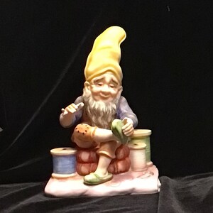 Peut inclure: Figurine en céramique d'un gnome réparant une chaussure. Le gnome porte un chapeau jaune, une chemise bleue et des chaussures vertes. Il est assis sur une pile de briques avec des bobines de fil à proximité. Design détaillé.