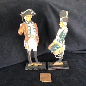 May include: Two vintage paper soldier figurines on wooden bases. The soldier on the left is wearing a red and white striped jacket and a tricorne hat. The soldier on the right is wearing a green and black uniform with a tall black hat. The bases are labeled "63rd Foot Officer 1768" and "43rd Foot Drum Major".
