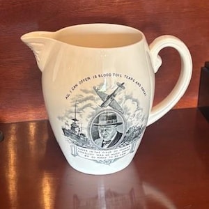 May include: Cream-colored ceramic pitcher with a handle and spout. The pitcher features a black and white illustration of a man in a hat, a ship, and an airplane. Text on the pitcher reads, "All I can offer is blood toil tears and sweat."