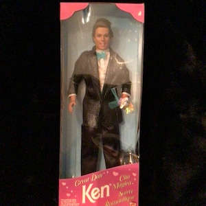 May include: A Ken doll wearing a black tuxedo with a teal bow tie and holding a pink rose. The box is pink and says "Great Date Ken Cita Magica Soiree Romantique".