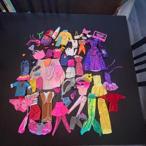 Op de afbeelding: Een grote verzameling miniatuur poppenkleding en accessoires, waaronder jurken, tops, broeken, rokken, een zeemeerminstaart, een roze speelgoedcassettespeler en diverse kleine items in diverse kleuren zoals paars, geel, roze, blauw en rood.