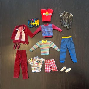 Op de afbeelding: Een verzameling poppenkleding bestaande uit een rood fluwelen jasje met wit overhemd, een rood fluwelen broek, een rood hemdje met afbeelding, een zwart glittervest, een blauwe broek met gele tailleband, een blauwe trui met patroon en een korte broek.