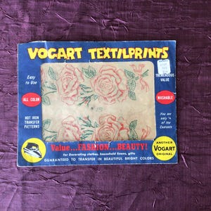Op de afbeelding: Een vintage verpakking met strijktransfers met een bloemenmotief met rozen. De verpakking is gelabeld met "VOCART TEXT/LPRINTS" en adverteert met "Value...FASHION...BEAUTY!" De verpakking is rood, wit en blauw.