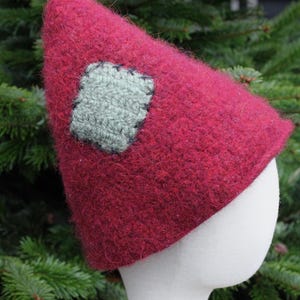 Peut inclure: Un chapeau en tricot bordeaux avec un patch en feutre vert cousu sur le devant. Le chapeau est en forme de cône et a une texture duveteuse.
