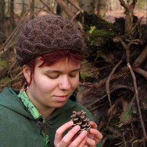 May include: A person wearing a brown knitted hat with a unique pattern holds a small pine cone in their hands. The background is a blurred image of a forest.