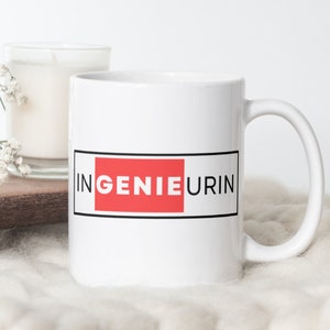May include: White ceramic mug with the word "INGENIEURIN" in black letters, with the word "GENIE" in a red rectangle. The mug has a white handle and is set against a soft, neutral background. A candle and flowers are in the background.