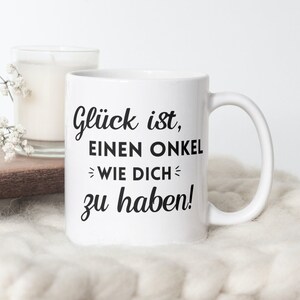 Puede incluir: Taza de cerámica blanca con texto negro que dice "Glück ist, einen Onkel wie dich zu haben!" La taza está sobre una superficie blanca y peluda.