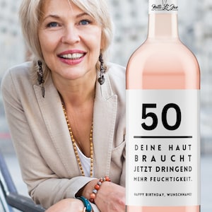 May include: A pink wine bottle with a white label that says "50" in black text. The label also says "Deine Haut Braucht Jetzt Dringend Mehr Feuchtigkeit." and "Happy Birthday, Wunschname!" in black text. The bottle is topped with a cork and has a white label with a black silhouette of a woman and the text "Netti Li Jae".