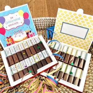 May include: Two boxes of chocolate bars with colorful wrappers. The boxes are open and the chocolate bars are visible. The boxes are decorated with a birthday theme and have a yellow and white pattern. The boxes are sitting on a woven basket.
