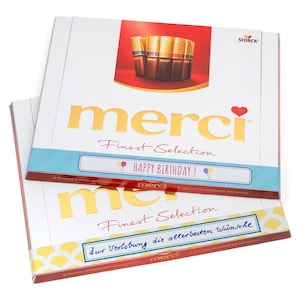 May include: A box of Merci Finest Selection chocolates with a gold foil design and a red heart. The box has a blue and white label with the text "Happy Birthday!" and a smaller label with the text "Zur Verlobung die allerbesten W&uuml;nsche".