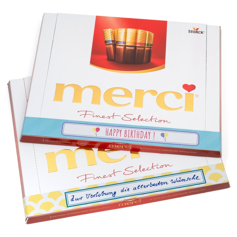 May include: A box of Merci Finest Selection chocolates with a gold foil design and a red heart. The box has a blue and white label with the text "Happy Birthday!" and a smaller label with the text "Zur Verlobung die allerbesten W&uuml;nsche".