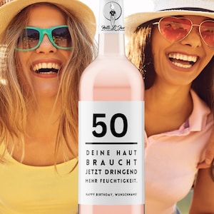 May include: A pink wine bottle with a white label that says "50" in black. The label also says "Deine Haut Braucht Jetzt Dringend Mehr Feuchtigkeit. Happy Birthday, Wunschaname!" in black text.