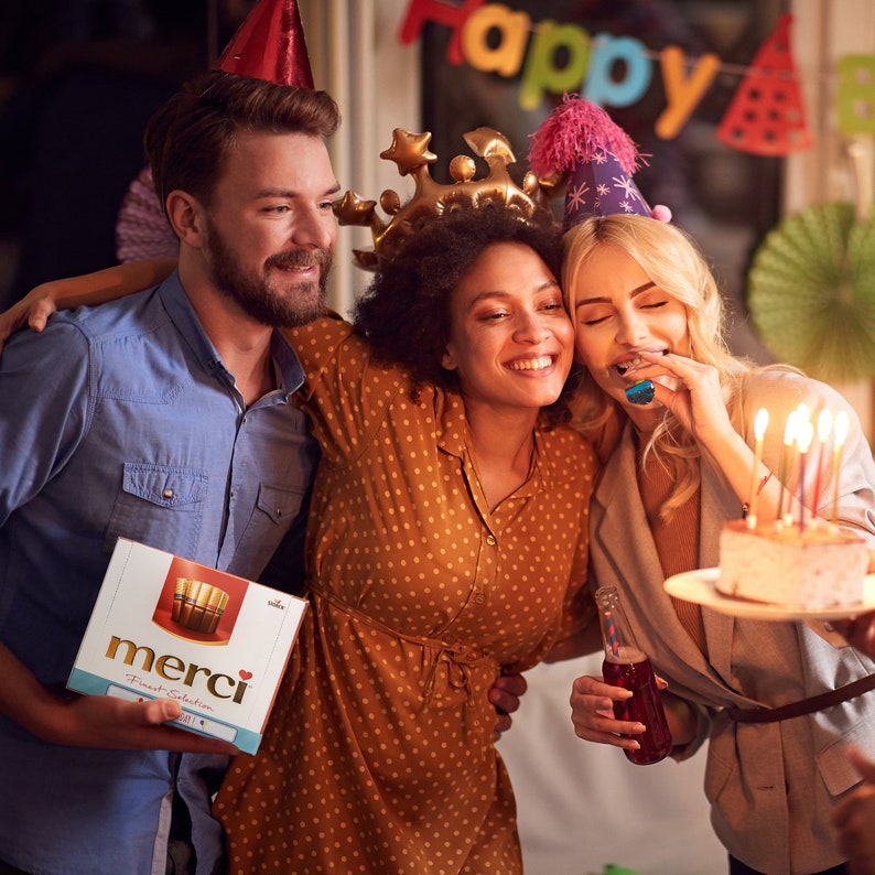 May include: Three friends celebrate a birthday with a cake and candles. One person holds a box of Merci Fine Selection chocolates. The box is red, white, and gold with the word "Merci" in a gold script font.