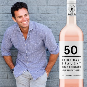May include: A pink wine bottle with a white label that says "50" and "DEINE HAUT BRAUCHT JETZT DRINGEND MEHR FEUCHTIGKEIT. HAPPY BIRTHDAY, WUNSCHNAME!"