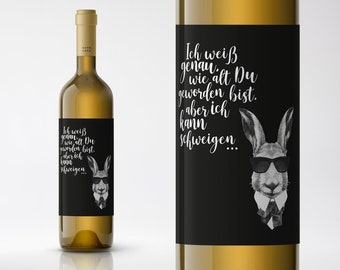 May include: A white wine bottle with a black label featuring a cartoon rabbit wearing sunglasses and a bow tie. The label text reads "Ich weiß genau wie alt Du geworden bist. aber ich kann schweigen..."