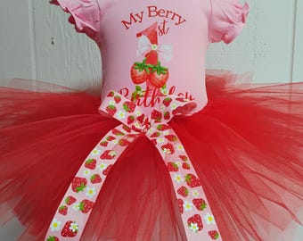 May include: A pink baby outfit with a red tutu skirt and a matching bow. The top says "My Berry 1st Birthday" with strawberry graphics. The tutu is layered with red and white tulle, and the bow features a strawberry pattern.