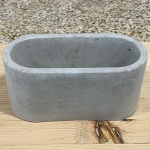 May include: A gray concrete oval planter with a smooth, slightly textured surface. The planter is ideal for small plants or succulents.
