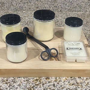 May include: Four glass candle jars with black lids and a white wax candle inside. A black candle wick trimmer and a small clear plastic container with a white label that reads "My So Happy Candle Co. Sugar Cookies 2.5 oz Wax Melts Hand Poured in Prosper, TX" are also on a wooden surface.