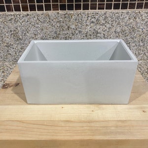 May include: A white rectangular concrete planter with a slightly angled top edge. The planter is sitting on a wooden surface.