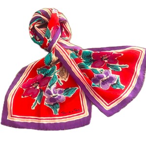 May include: A rectangular scarf featuring a vibrant red background with a floral pattern in shades of purple, magenta, and teal green. The scarf has a purple border with thin white and pink stripes and includes the signature text Oscar de la Renta.