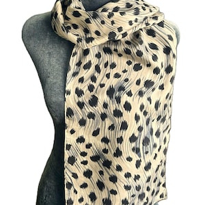 May include: A rectangular scarf featuring a beige background with a repeating pattern of black, irregular, elongated spots. The fabric displays subtle, wavy, vertical lines throughout the design, creating a textured visual effect across the surface.